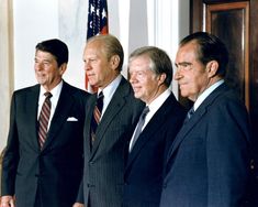 four men in suits standing next to each other with an american flag on the wall behind them