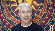 an older man with white hair standing in front of a colorful wall and painting behind him