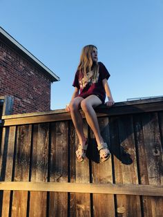 a young woman sitting on top of a wooden fence next to a brick building and looking off into the distance