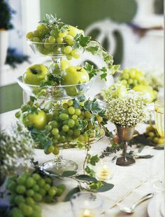 a table topped with lots of fruit and greenery next to a vase filled with green apples