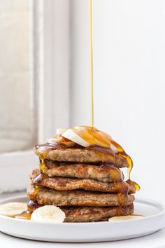 a stack of pancakes with syrup and bananas on a white plate next to a window