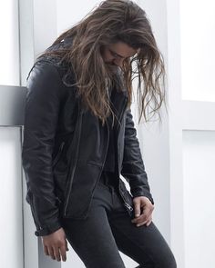 a man with long hair wearing a black leather jacket and jeans leaning against a wall