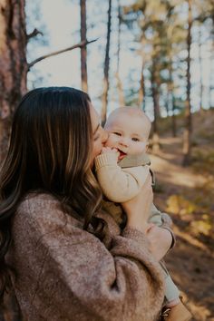 a woman holding a baby in her arms and smiling at the camera with trees behind her