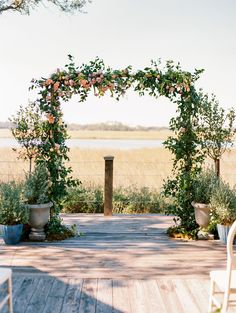 a wooden deck with chairs and an arch covered in greenery next to a body of water