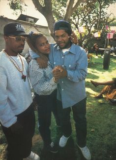 two men and a woman standing next to each other in front of a house with trees