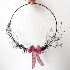 a hand holding up a black wire wreath with pink ribbon and leaves on it, against a white wall