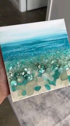 a hand holding up a piece of art that looks like sea glass and rocks on the beach