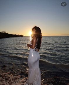 a woman in white dress standing on the beach near water at sun set with her back turned to the camera