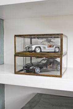 two model cars in a glass case sitting on top of a white shelf next to each other