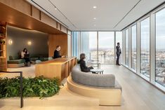 people are sitting and standing in an office with floor to ceiling windows overlooking the city