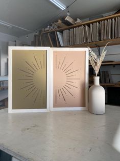 two framed pictures sitting on top of a counter next to a vase and bookshelf