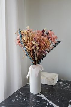 a bouquet of dried flowers in a white vase on a black marble table next to a window