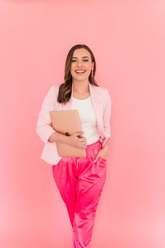 a woman in pink pants and white shirt holding a folder while standing against a pink background