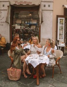 three women sitting at an outdoor table talking and drinking wine in front of a store