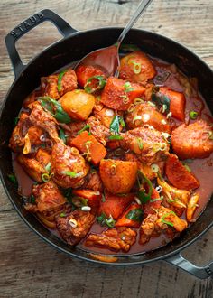 a skillet filled with meat and vegetables on top of a wooden table next to a spoon