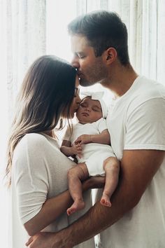 a man and woman kissing while holding a baby in front of a window with curtains