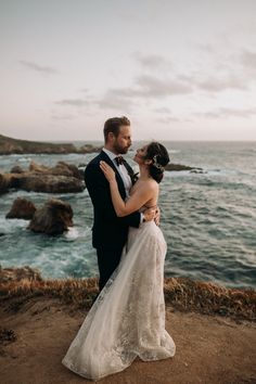 a bride and groom standing on the cliff by the ocean at sunset with their arms around each other