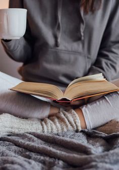 a person sitting on a bed reading a book and holding a coffee cup in their hands