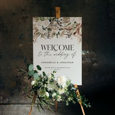 a welcome sign with flowers and greenery is displayed in front of an easel