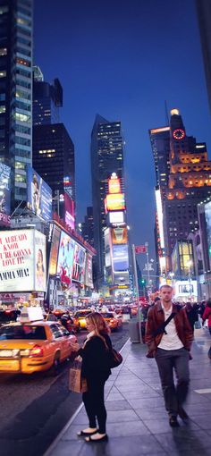 people are walking on the sidewalk in times square at night with billboards and taxi cabs