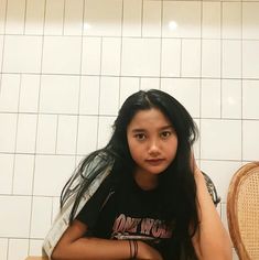 a young woman sitting on top of a wooden chair next to a white tiled wall