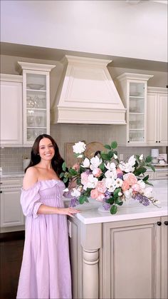 a woman standing in front of a kitchen island with flowers on the counter and cabinets