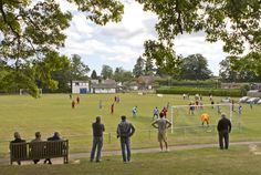 several people are standing on the grass watching a soccer game from behind a wire fence