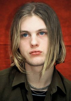 a man with long blonde hair and blue eyes wearing a green shirt looking at the camera