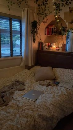 an unmade bed with a laptop on it in front of a window and bookshelf