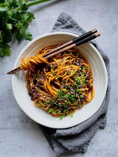 a white bowl filled with noodles and vegetables next to chopsticks on a gray surface