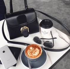 a cup of coffee, cell phone and purse on a white table with a black handbag