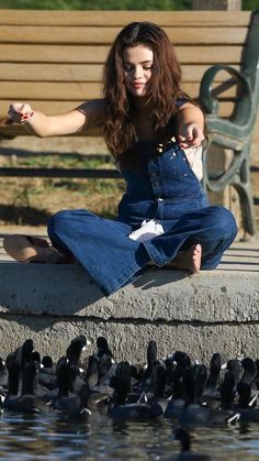 a woman is sitting on the edge of a pond with ducks in front of her