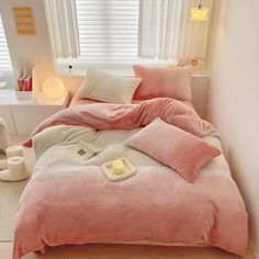 a bed with pink comforter and pillows in a white room next to a window