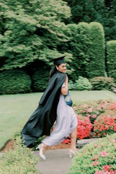 a woman in a graduation gown and cap is running through the garden with flowers on either side