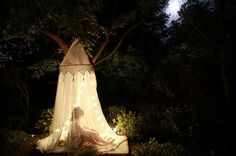 a woman sitting in the middle of a forest at night under a canopy with fairy lights on it