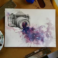 a watercolor painting of a camera on paper next to paintbrushes and other art supplies