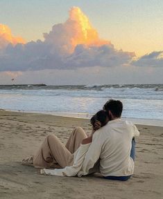 a man and woman sitting on the beach watching the sun go down over the ocean