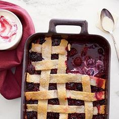 a berry pie in a baking dish next to a bowl of whipped cream and spoons