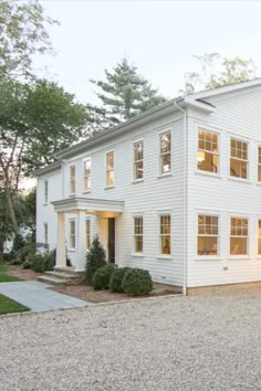 a large white house sitting on top of a gravel lot next to trees and bushes