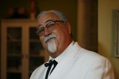 an older man wearing glasses and a white suit with a black tie is looking at the camera