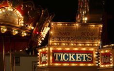 an illuminated ticket booth at night time with lights on it's sides and the word tickets written in large letters