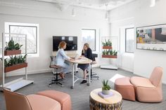 two women are sitting at a table in an office with pink chairs and potted plants