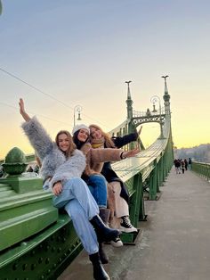 three women sitting on the edge of a bridge taking a selfie with their arms in the air