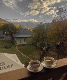 two cups of coffee sit on a wooden table overlooking a mountain valley with houses in the distance