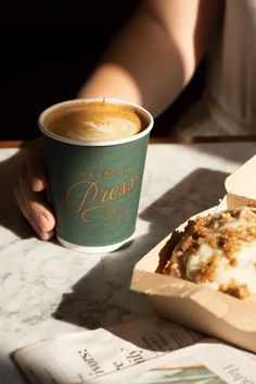 a person holding a cup of coffee next to a pastry in a box on a table