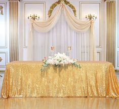 the table is covered with gold sequins and white flowers in front of a golden drape