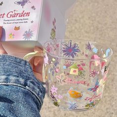 a person holding a glass cup with flowers on it and a book in the background