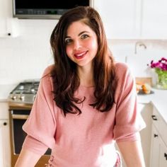 a woman standing in a kitchen next to a counter with food on it and smiling at the camera