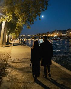 two people walking down a sidewalk next to the water at night with full moon in background