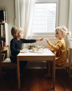 two young children sitting at a table with cups and saucers on it, one holding the other's hand